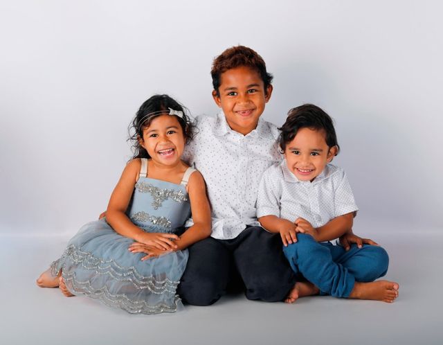 Three siblings sitting close on a grey backdrop: a girl in a sparkly blue dress and two boys in white shirts, all smiling
