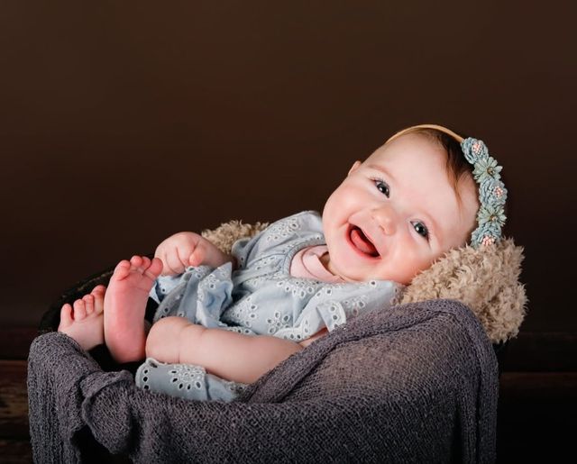 A laughing baby in a blue eyelet outfit and flower headband, lying back in a wrapped basket
